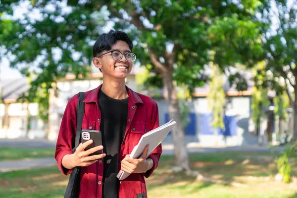 ハッピーな表情でスマートフォンを使用したアジアの若い大学生 公立公園で携帯と本を持っている間 男性は笑顔で コピースペース — ストック写真