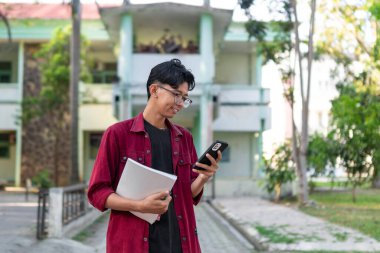 Genç Asyalı üniversite öğrencisi mutlu bir ifadeyle akıllı telefon kullanıyor. Halk parkında elinde telefon ve kitaplarla gülümseyen bir erkek. Boşluğu kopyala