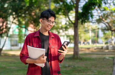 Genç Asyalı üniversite öğrencisi mutlu bir ifadeyle akıllı telefon kullanıyor. Halk parkında elinde telefon ve kitaplarla gülümseyen bir erkek. Boşluğu kopyala