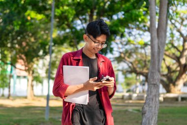 Genç Asyalı üniversite öğrencisi mutlu bir ifadeyle akıllı telefon kullanıyor. Halk parkında elinde telefon ve kitaplarla gülümseyen bir erkek. Boşluğu kopyala