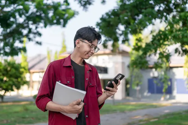 Genç Asyalı üniversite öğrencisi mutlu bir ifadeyle akıllı telefon kullanıyor. Halk parkında elinde telefon ve kitaplarla gülümseyen bir erkek. Boşluğu kopyala