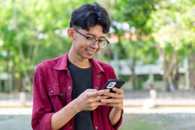 Telefonunu tutarken gülümseyen Asyalı bir adamın portresi. Kırmızı gömlekli adam akıllı telefon kullanırken kameraya neşeyle gülümsüyor.