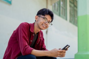 Telefonunu tutarken gülümseyen Asyalı bir adamın portresi. Kırmızı gömlekli adam akıllı telefon kullanırken kameraya neşeyle gülümsüyor.