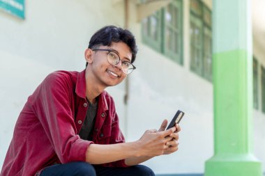 Telefonunu tutarken gülümseyen Asyalı bir adamın portresi. Kırmızı gömlekli adam akıllı telefon kullanırken kameraya neşeyle gülümsüyor.