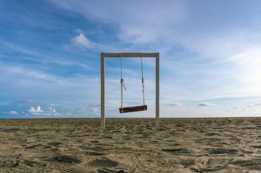 Parlak mavi bir gökyüzünün altında kumlu bir sahilde tek başına duran ahşap bir salıncak..