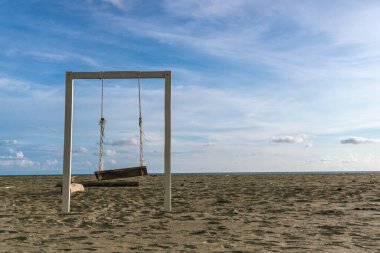 Parlak mavi bir gökyüzünün altında kumlu bir sahilde tek başına duran ahşap bir salıncak..