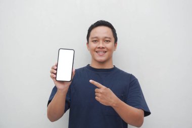 Genç Asyalı adam gülümsüyor ve beyaz arka planda izole edilmiş akıllı telefon ekranına işaret ediyor. Yaşam tarzı ve reklam konsepti. Mavi gömlekli adamlar ellerinde telefon modeli tutuyorlar.