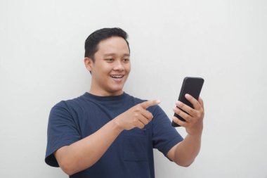 Genç Asyalı adam gülümsüyor ve beyaz arka planda izole edilmiş akıllı telefona işaret ediyor. Yaşam tarzı ve reklam konsepti. Telefon tutan mavi gömlekli adamlar.