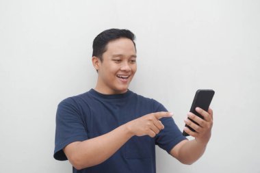 Genç Asyalı adam gülümsüyor ve beyaz arka planda izole edilmiş akıllı telefona işaret ediyor. Yaşam tarzı ve reklam konsepti. Telefon tutan mavi gömlekli adamlar.