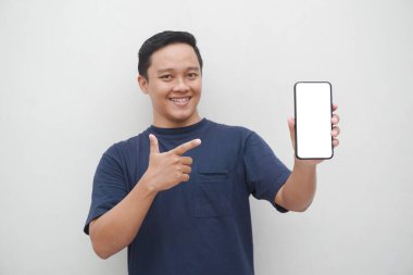 Genç Asyalı adam gülümsüyor ve beyaz arka planda izole edilmiş akıllı telefon ekranına işaret ediyor. Yaşam tarzı ve reklam konsepti. Mavi gömlekli adamlar ellerinde telefon modeli tutuyorlar.