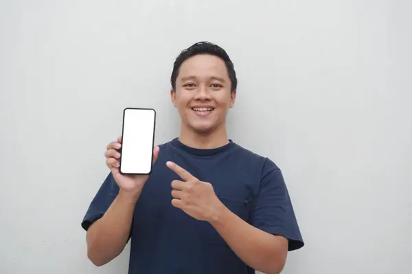 Genç Asyalı adam gülümsüyor ve beyaz arka planda izole edilmiş akıllı telefon ekranına işaret ediyor. Yaşam tarzı ve reklam konsepti. Mavi gömlekli adamlar ellerinde telefon modeli tutuyorlar.
