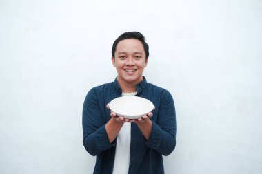 Elinde mutlu ve heyecanlı bir ifadeyle boş bir tabak tutarken mavi gömlek giyen mutlu Asyalı bir adamın portresi. Genç adam boş model plakayı gösteriyor ve işaret ediyor. Beyaz arkaplan ile izole edilmiş.