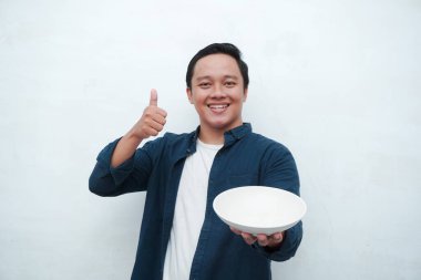 Elinde mutlu ve heyecanlı bir ifadeyle boş bir tabak tutarken mavi gömlek giyen mutlu Asyalı bir adamın portresi. Genç adam boş model plakayı gösteriyor ve işaret ediyor. Beyaz arkaplan ile izole edilmiş.