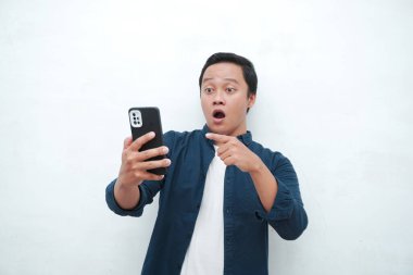 Genç Asyalı adam şok olmuş ve şaşırmış bir ifadeyle akıllı telefona benziyor. Mavi tişörtlü erkek, beyaz arkaplanda izole edilmiş heyecanlı bir ifade sergiliyor