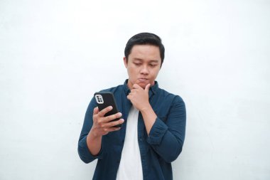 Genç Asyalı adam cep telefonu tutarken düşünüp konsantre oluyor. Beyaz arkaplan üzerinde telefon tutarken Asyalı öğrenci düşünüyor ve fikir arıyor
