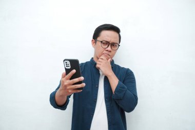 Genç Asyalı adam cep telefonu tutarken düşünüp konsantre oluyor. Beyaz arkaplan üzerinde telefon tutarken Asyalı öğrenci düşünüyor ve fikir arıyor