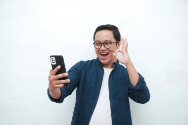 Genç Asyalı adam şok olmuş ve şaşırmış bir ifadeyle akıllı telefona benziyor. Mavi tişörtlü erkek, beyaz arkaplanda izole edilmiş heyecanlı bir ifade sergiliyor