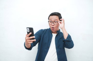 Genç Asyalı adam şok olmuş ve şaşırmış bir ifadeyle akıllı telefona benziyor. Mavi tişörtlü erkek, beyaz arkaplanda izole edilmiş heyecanlı bir ifade sergiliyor