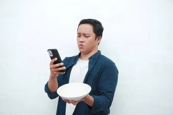 Ne yiyeceğini düşünürken telefonu tutan kafası karışmış Asyalı bir adamın portresi. Genç adam elinde boş bir maket tabağı tutarken telefondan yemek sipariş ediyor. Beyaz arkaplan ile izole edilmiş.