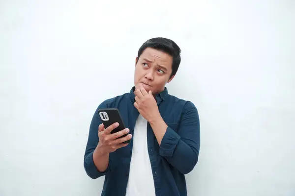 Genç Asyalı adam cep telefonu tutarken düşünüp konsantre oluyor. Beyaz arkaplan üzerinde telefon tutarken Asyalı öğrenci düşünüyor ve fikir arıyor