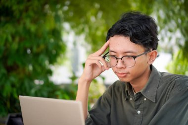 Genç Asyalı adam bilgisayarına bakarken düşünüyor. Sabahları açık kafede dizüstü bilgisayarıyla çalışan bir adam.