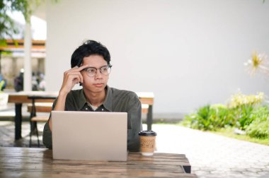 Genç Asyalı adam bilgisayarına bakarken düşünüyor. Sabahları açık kafede dizüstü bilgisayarıyla çalışan bir adam.