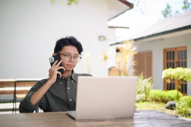 Genç Asyalı adam cep telefonuyla konuşurken dizüstü bilgisayarıyla çalışıyor. Mavi gömlekli bir erkek çalışırken telefonla konuşuyor.