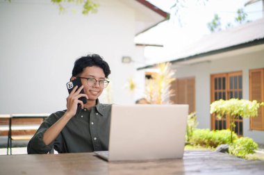 Genç Asyalı adam cep telefonuyla konuşurken dizüstü bilgisayarıyla çalışıyor. Mavi gömlekli bir erkek çalışırken telefonla konuşuyor.