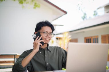 Genç Asyalı adam cep telefonuyla konuşurken dizüstü bilgisayarıyla çalışıyor. Mavi gömlekli bir erkek çalışırken telefonla konuşuyor.