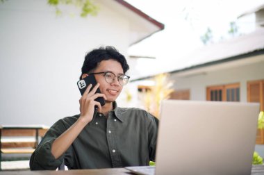 Genç Asyalı adam cep telefonuyla konuşurken dizüstü bilgisayarıyla çalışıyor. Mavi gömlekli bir erkek çalışırken telefonla konuşuyor.