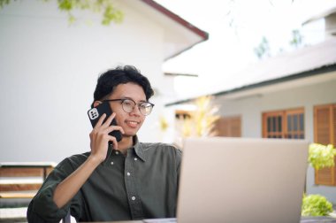 Genç Asyalı adam cep telefonuyla konuşurken dizüstü bilgisayarıyla çalışıyor. Mavi gömlekli bir erkek çalışırken telefonla konuşuyor.