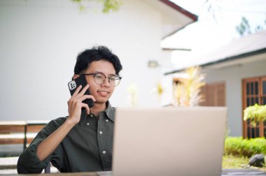 Genç Asyalı adam cep telefonuyla konuşurken dizüstü bilgisayarıyla çalışıyor. Mavi gömlekli bir erkek çalışırken telefonla konuşuyor.
