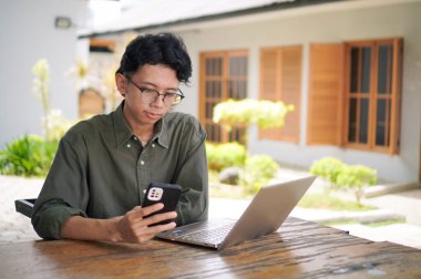 Dizüstü bilgisayarı üzerinde çalışırken elinde akıllı telefon tutan adam. Asyalı erkek, açık kafedeki telefonuna ve bilgisayarına odaklanmış.