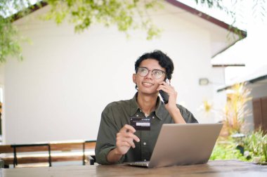 Cep telefonuyla konuşan ve elinde kredi kartı tutan Asyalı genç bir adam dizüstü bilgisayarıyla çalışıyor. Çevrimiçi satın almaya çalışan adam kartla