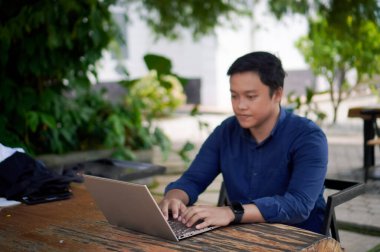 Mavi gömlekli bir adam dışarıdaki kafede laptopuyla çalışıyor.