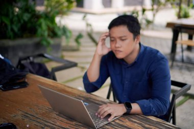 Asyalı bir adam dizüstü bilgisayarla uğraşırken stres ve yorgunluk yüzünden başını tutuyor. Asyalı işadamının migreni ya da iş bitiminden kaynaklanan baş ağrısı var