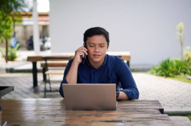 Genç Asyalı adam cep telefonuyla konuşurken dizüstü bilgisayarıyla çalışıyor. Mavi gömlekli bir erkek çalışırken telefonla konuşuyor.