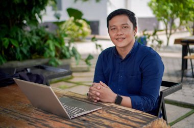 Kameraya gülümseyen mavi gömlekli genç Asyalı adam. Adam dizüstü bilgisayarıyla çalışırken pozitif enerji gösteriyor..