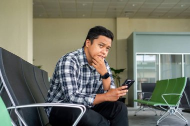 Male Tourist Sitting in the Airport waiting for plane.Asian man thingking while using mobile phone in airport waiting area. clipart