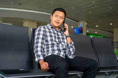 Male Tourist calling with his phone while waiting for departure in the airport. Asian man using mobile phone in airport waiting area. clipart