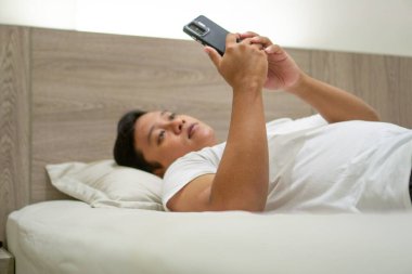 Geceleri yatakta akıllı telefon kullanan bir adam. Genç Asyalı adam akıllı telefon kullanırken yatakta yatıyor. Kötü alışkanlık.
