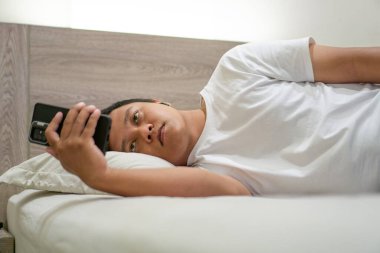 Geceleri yatakta akıllı telefon kullanan bir adam. Genç Asyalı adam akıllı telefon kullanırken yatakta yatıyor. Kötü alışkanlık.