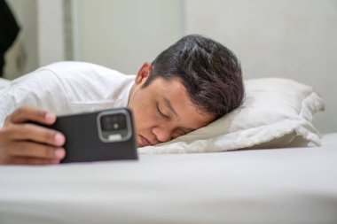 Geceleri yatakta uyuyan ve hala telefonunu tutan bir adam. Kötü alışkanlık. Yorgun Asyalı adam cep telefonu kullanırken yatakta uyuyakalıyor. Kötü alışkanlık.