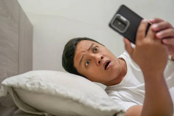 Adam gece yatakta akıllı telefon tutarken şaşırmış bir ifade sergiliyor. Genç Asyalı adam şok olmuş bir ifadeyle akıllı telefon kullanırken yatakta yatıyor..