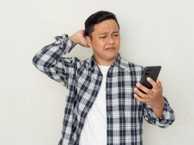 Genç Asyalı adam cep telefonuyla düşünürken kafası karışıyor. Asyalı bir öğrenci kafasını kaşıyarak beyaz arka plan üzerinden telefonuna bakıyor.