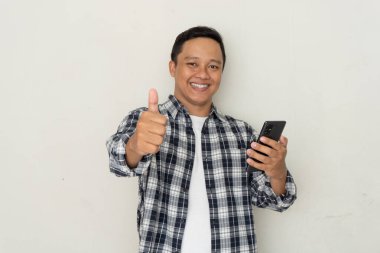 Genç Asyalı adam mutlu gülümsüyor ve cep telefonu tutarken başparmak işareti yapıyor..