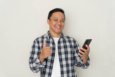 Genç Asyalı adam mutlu gülümsüyor ve cep telefonu tutarken başparmak işareti yapıyor..