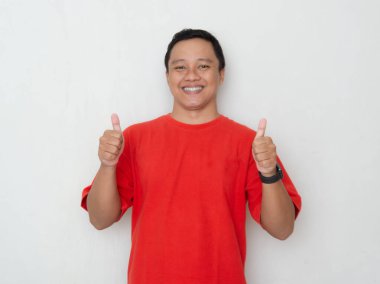 Kırmızı tişörtlü genç Asyalı adam beyaz arka planda güvenle gülümsüyor. Pozitiflik, onay ve memnuniyet kavramı
