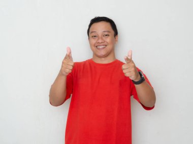 Kırmızı tişörtlü genç Asyalı adam beyaz arka planda güvenle gülümsüyor. Fotoğraf pozitiflik, onay ve memnuniyet kavramını gösterir