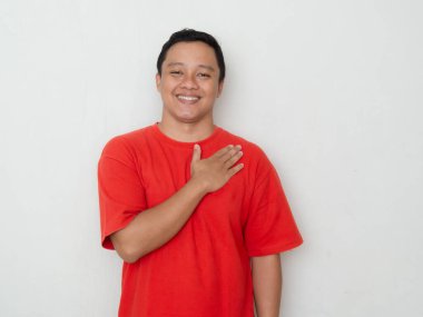 Smiling young Asian man in red shirt with hand over heart, expressing gratitude or sincerity. concept of warmth, thankfulness, and genuine emotion. clipart